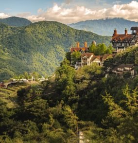 An aerial shot of a small village on the hill surrounded by forested mountains